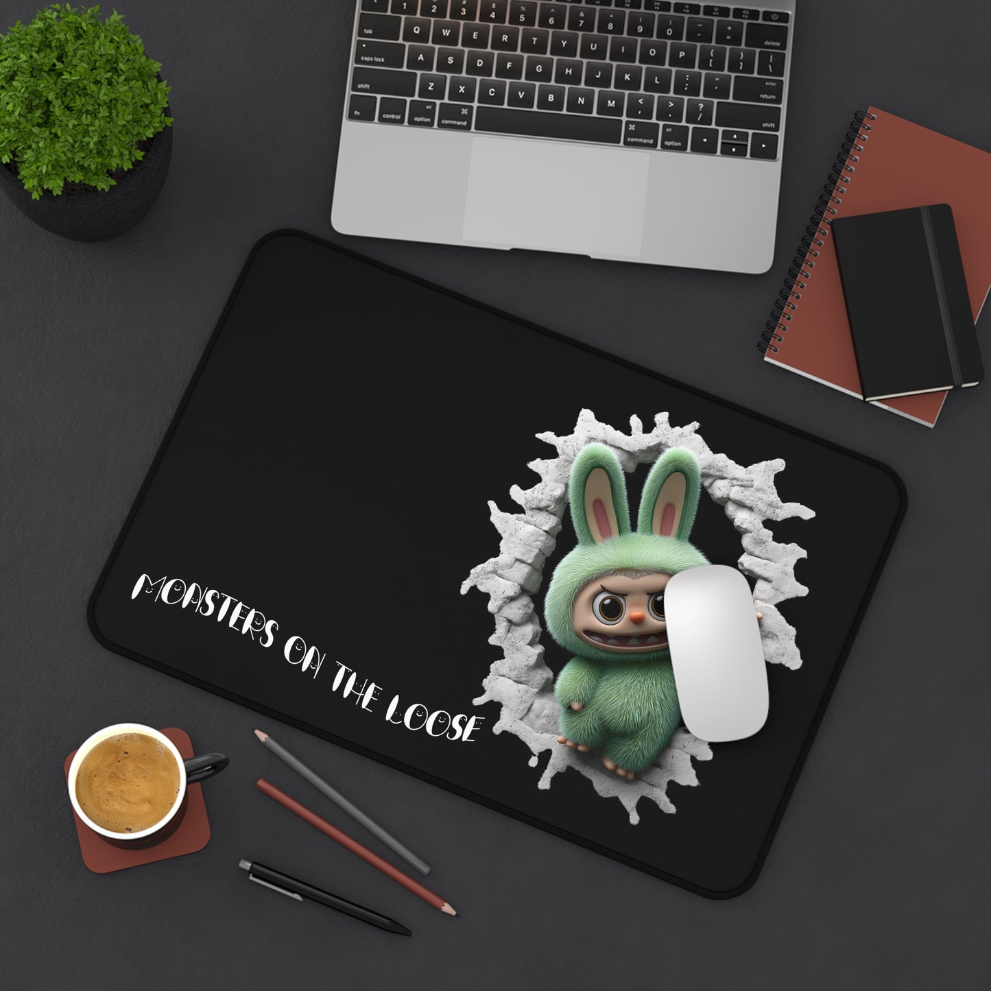 Desk setup with a black mouse pad featuring a green creature design and text, next to a laptop and office supplies.