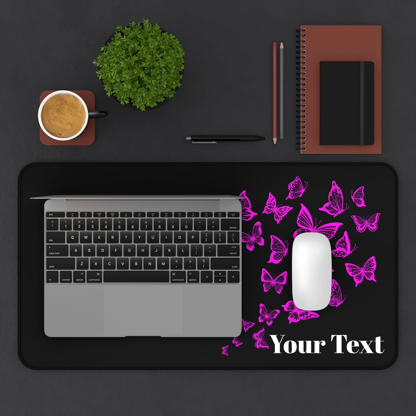 Black desk mat with pink butterflies and 'Your Text' placeholder, placed on a dark surface with a laptop, mouse, and office items.