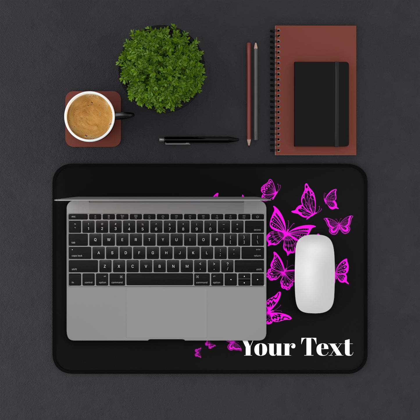 Laptop on a black mouse pad with pink butterflies and 'Your Text' placeholder, surrounded by office supplies on a dark surface.