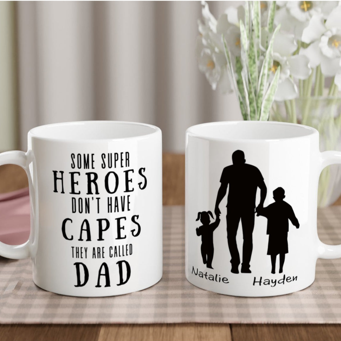 Two glossy white ceramic mugs with personalized text and a graphic design, one featuring a superhero illustration and the other with a more abstract design, both with a dad and children figure.