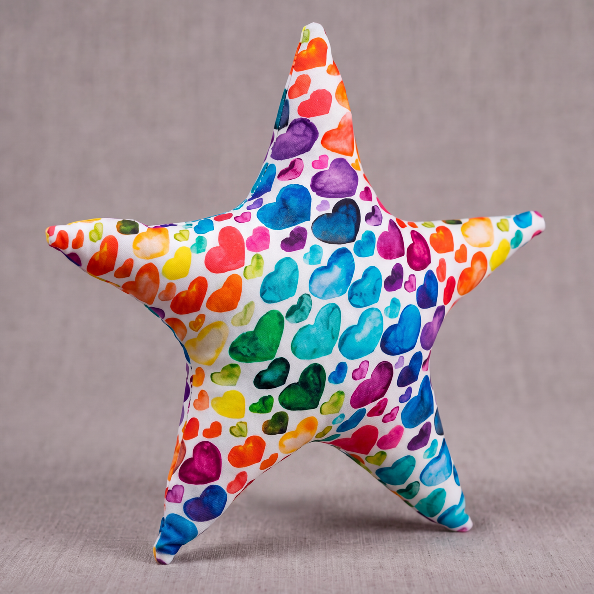 A five-pointed star plush toy covered in a colorful watercolor heart pattern, set against a neutral gray textured background with text at the bottom reading 'THE RAINBOW HEART STAR Watercolor Pattern Textile Plush'.