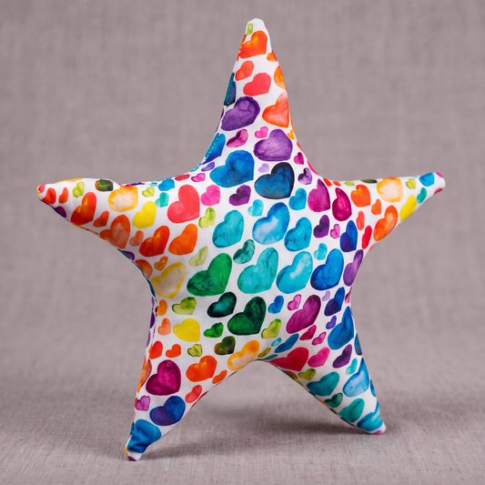 A five-pointed star plush toy covered in a colorful watercolor heart pattern, set against a neutral gray textured background with text at the bottom reading 'THE RAINBOW HEART STAR Watercolor Pattern Textile Plush'.