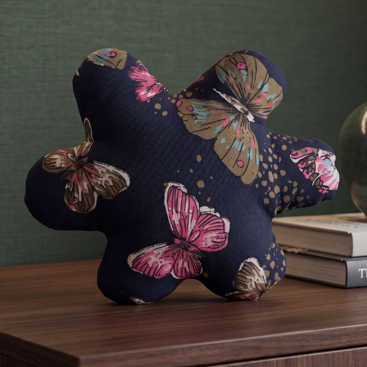 A star-shaped plush cushion with a navy blue fabric printed with pink, brown, and gold butterflies, styled on a wooden table next to books against a dark green textured wall.