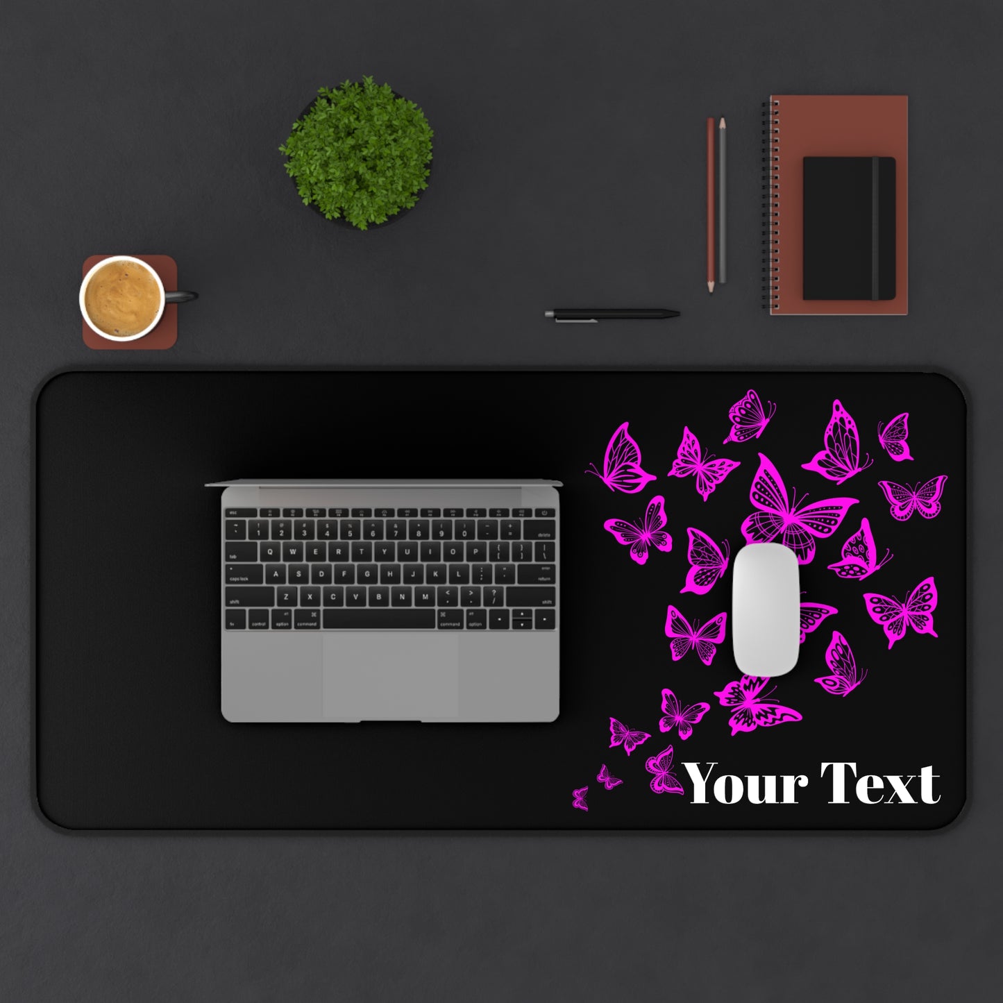 Black desk mat with pink butterflies and 'Your Text' placeholder, placed on a dark surface with laptop, mouse, and stationery items.