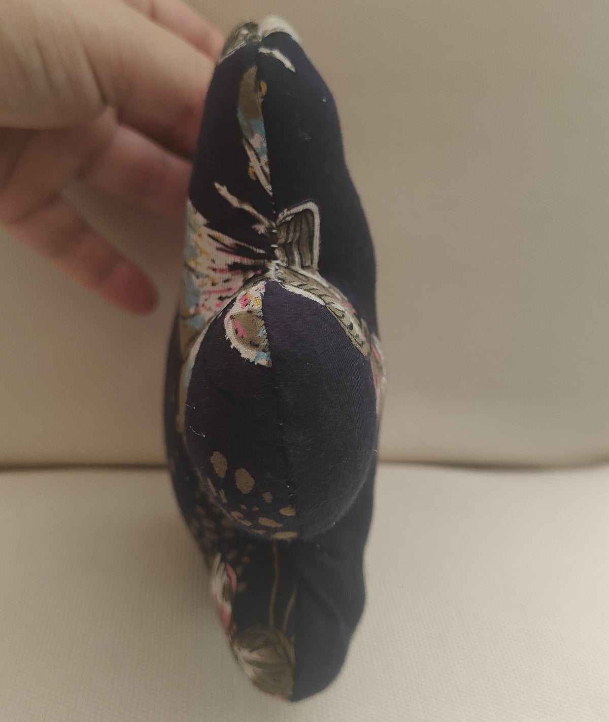 A hand holding a small decorative cushion or pillow made of dark navy fabric with a multicolor floral pattern, set against a plain beige background.