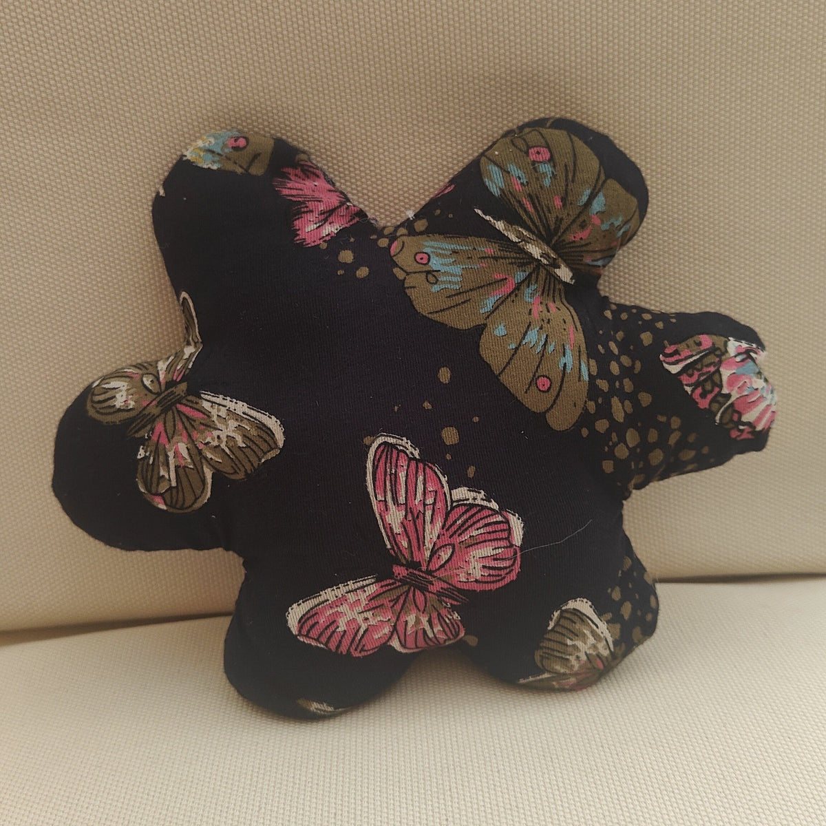 A star-shaped plush cushion or pillow with a black fabric base printed with colorful butterfly and gold polka dot patterns, resting on a beige textured fabric surface.