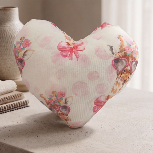 A heart-shaped decorative pillow with a giraffe and pink bow print, resting on a light stone surface next to a neutral vase.