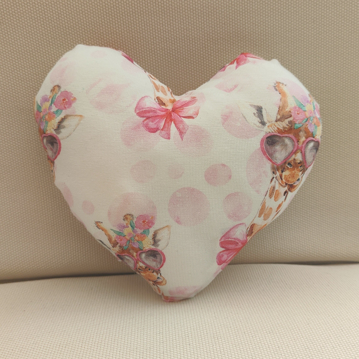 A heart-shaped decorative pillow with a white background, pink polka dots, and a watercolor floral print, resting on a beige textured fabric surface.