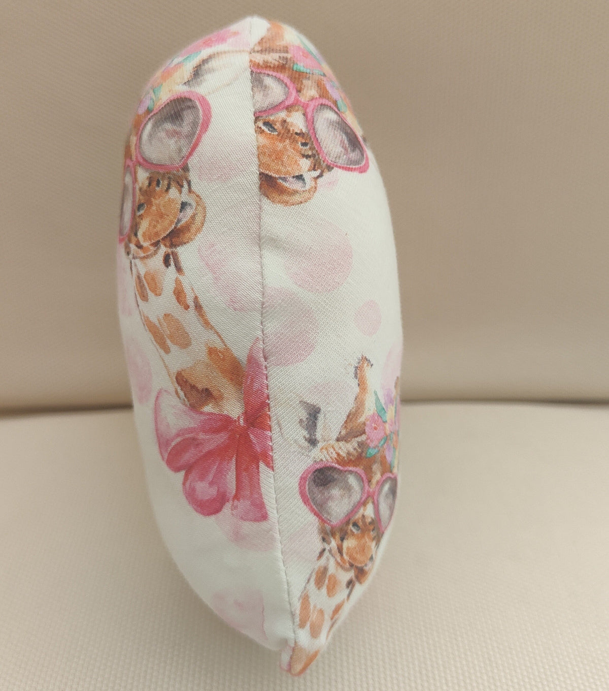 A small decorative cushion or pillow with a white background and a whimsical print of giraffes wearing pink sunglasses and large pink bows, standing upright against a neutral beige backdrop.