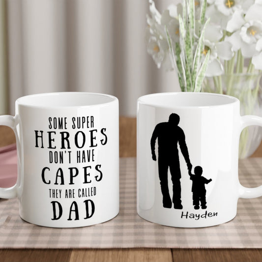 Two ceramic mugs with personalized text, one featuring an illustration of a dad and child and the other with text alone, placed on a table with a floral arrangement and a wooden background.