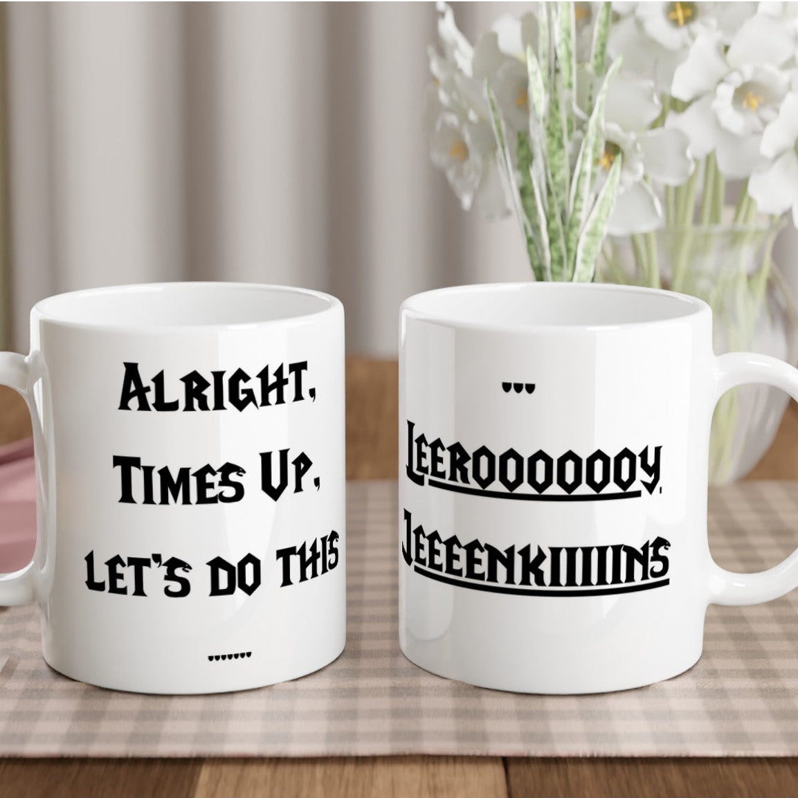 Two white mugs with humorous text on a table with a checkered tablecloth and flowers in the background.