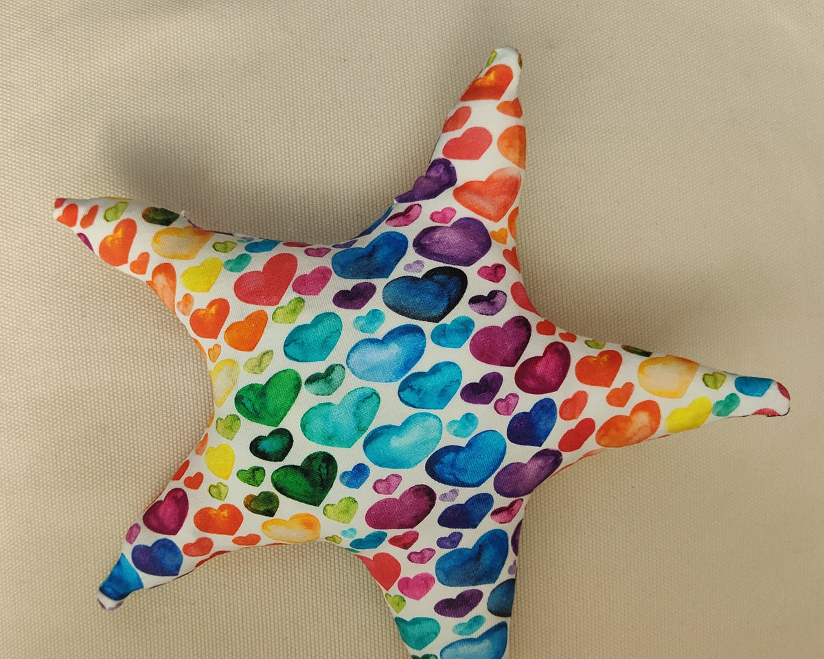 A starfish-shaped decorative pillow featuring a colorful watercolor heart pattern on a white background, displayed on a beige textured fabric surface.